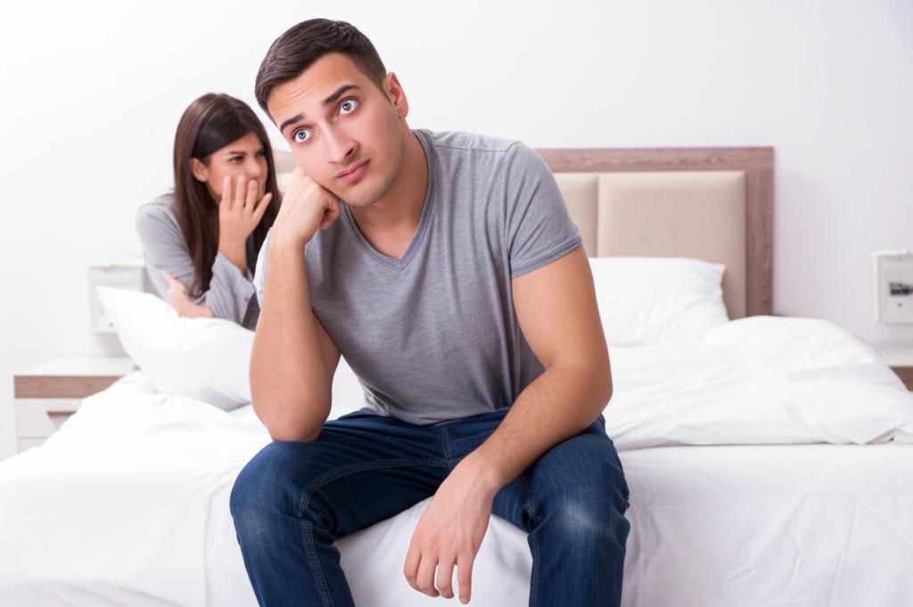 A tired man sitting on the edge of the bed, looking stressed and exhausted, with a concerned woman beside him.